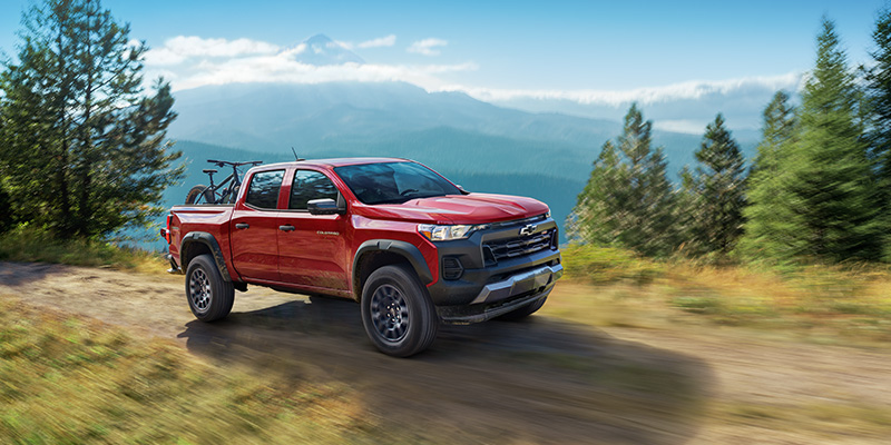 Red 2026 Chevy Colorado driving on a dirt trail through a forested mountain landscape